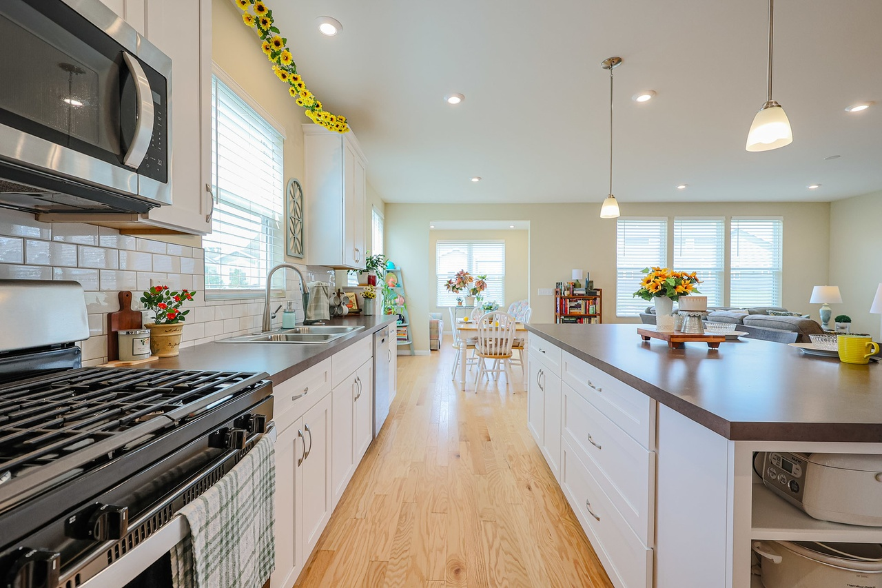 Bright kitchen with white cabinets, wooden countertops, a stove, microwave, and sink. Sunflower decorations and plants are on the window sills. Windows with white blinds let in natural light, and a dining area with a table and chairs is visible in th