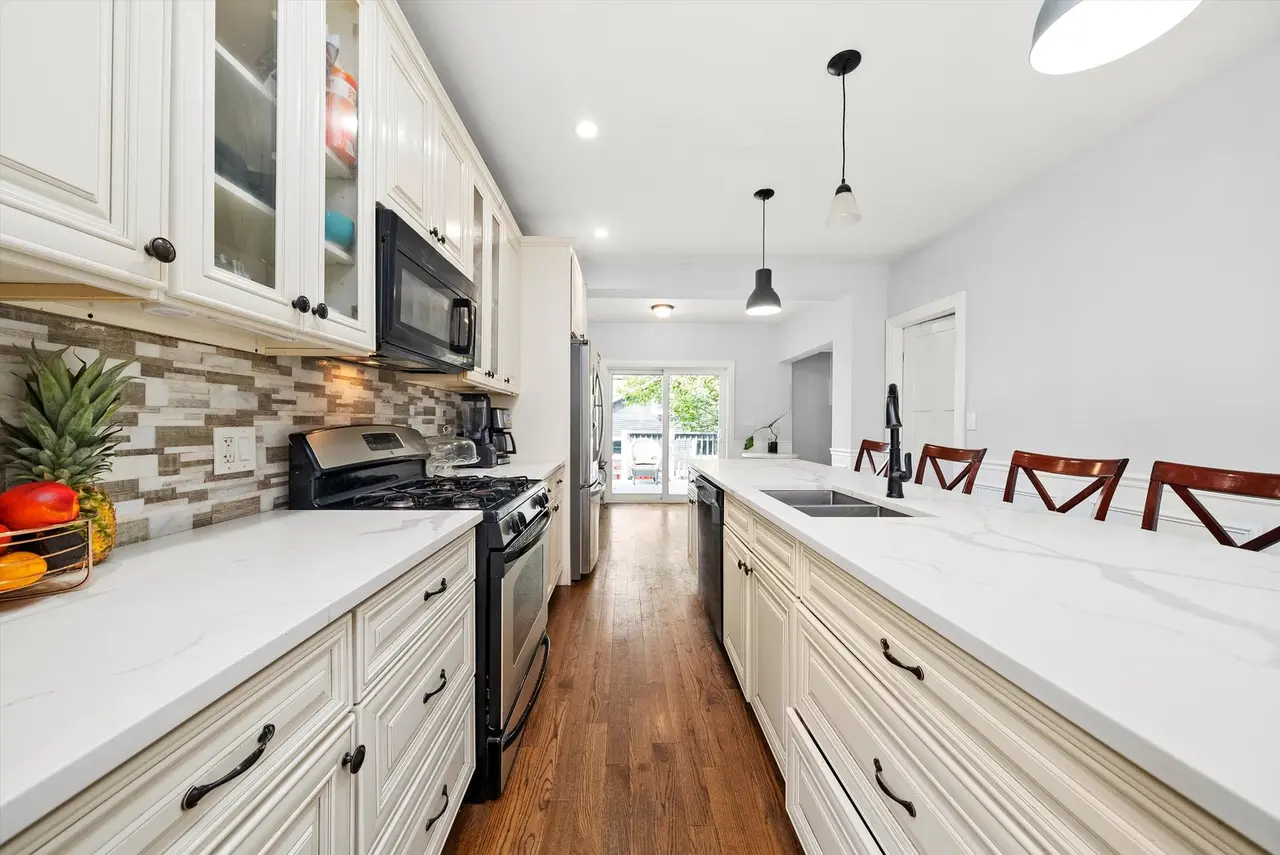 Modern kitchen with white cabinetry, black appliances, a white marble countertop, bar stools, hardwood floors, and a sliding glass door leading outside.
