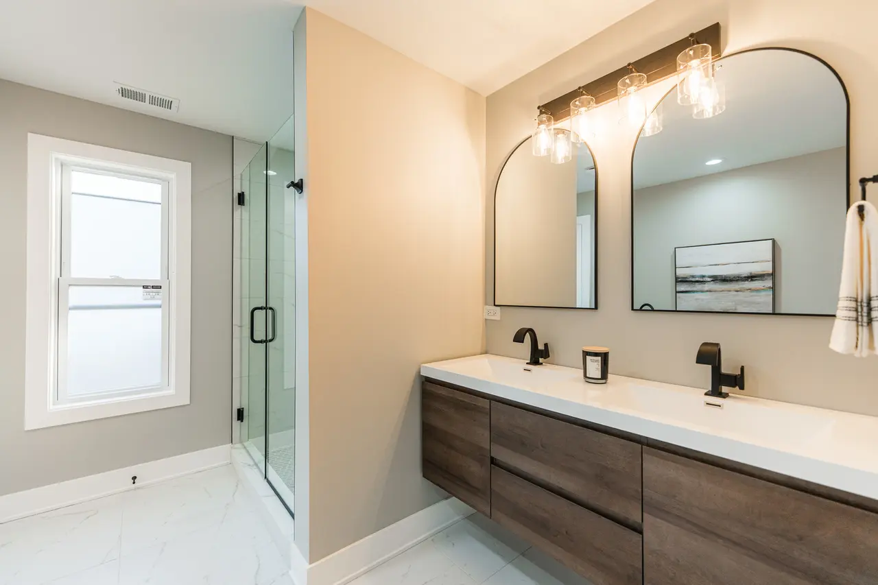 Modern bathroom with a double sink vanity, large mirrors, wall-mounted black faucets, and a glass-enclosed shower with a window.