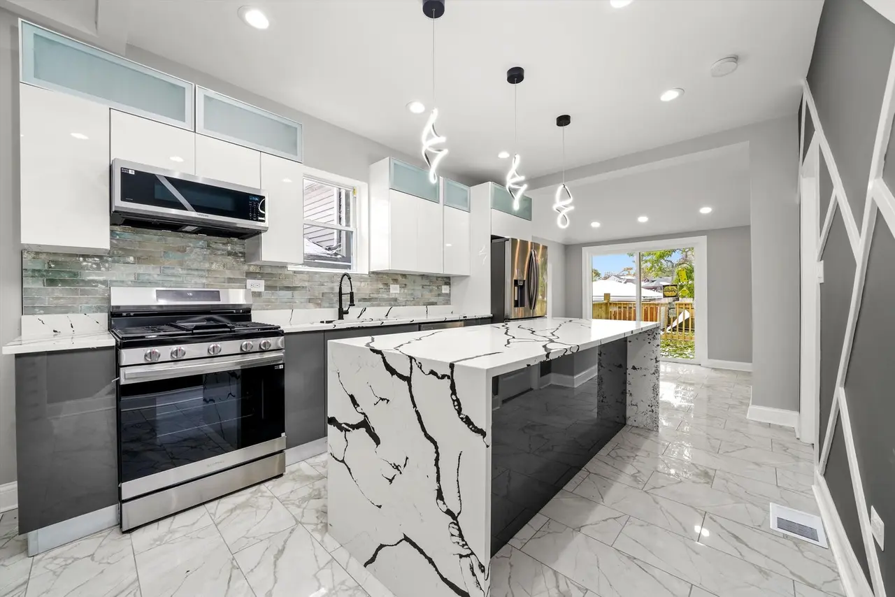 Modern kitchen with white cabinets, stainless steel stove, black and white marble island, glass backsplash, and gray walls. There are pendant lights above the island and a sliding glass door leading outside.