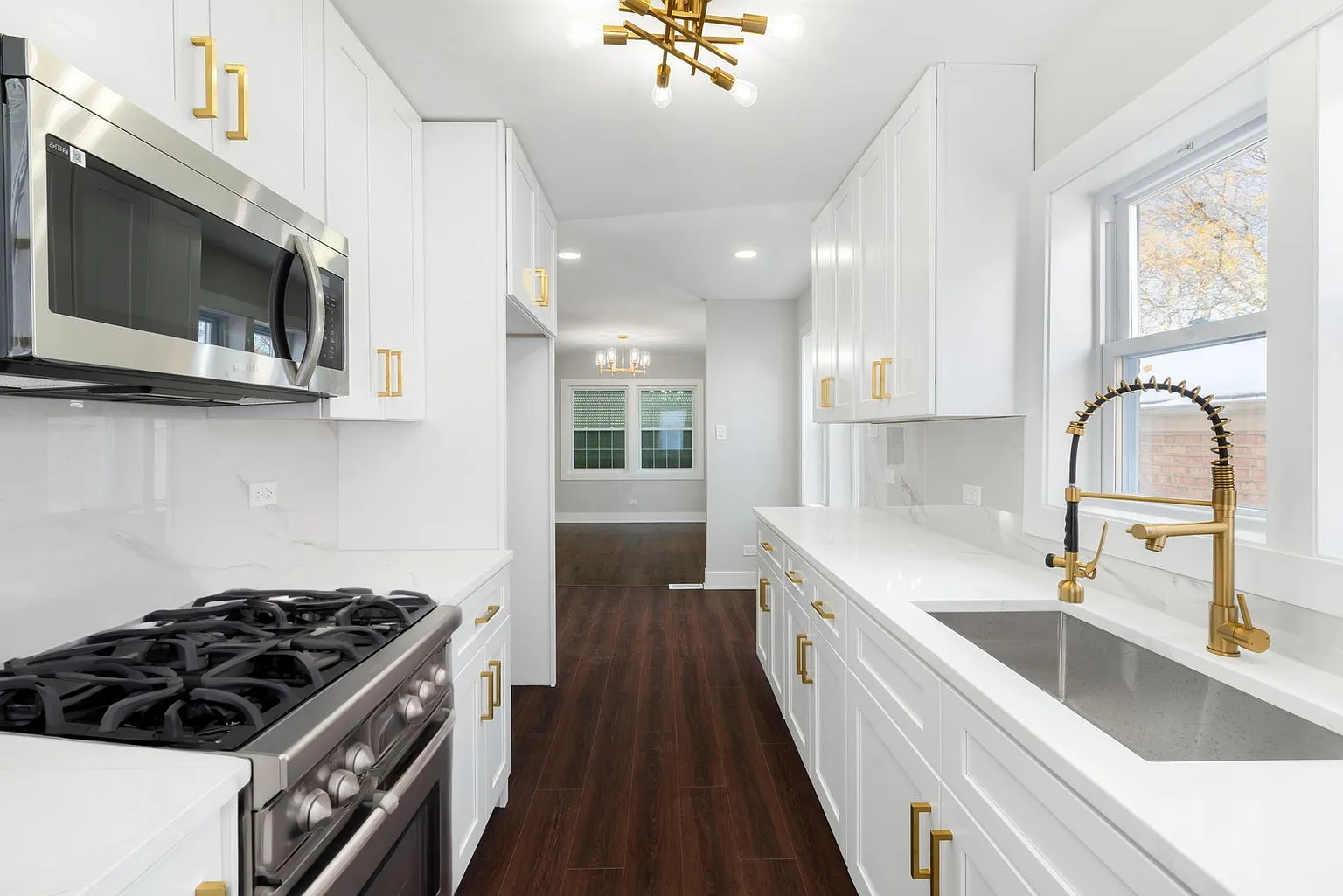 Modern kitchen with white cabinets, gold handles, stainless steel appliances, a white countertop with beige veining, a farmhouse sink with a gold and black faucet, and dark hardwood floors. There are two windows above the sink and a modern gold ceili