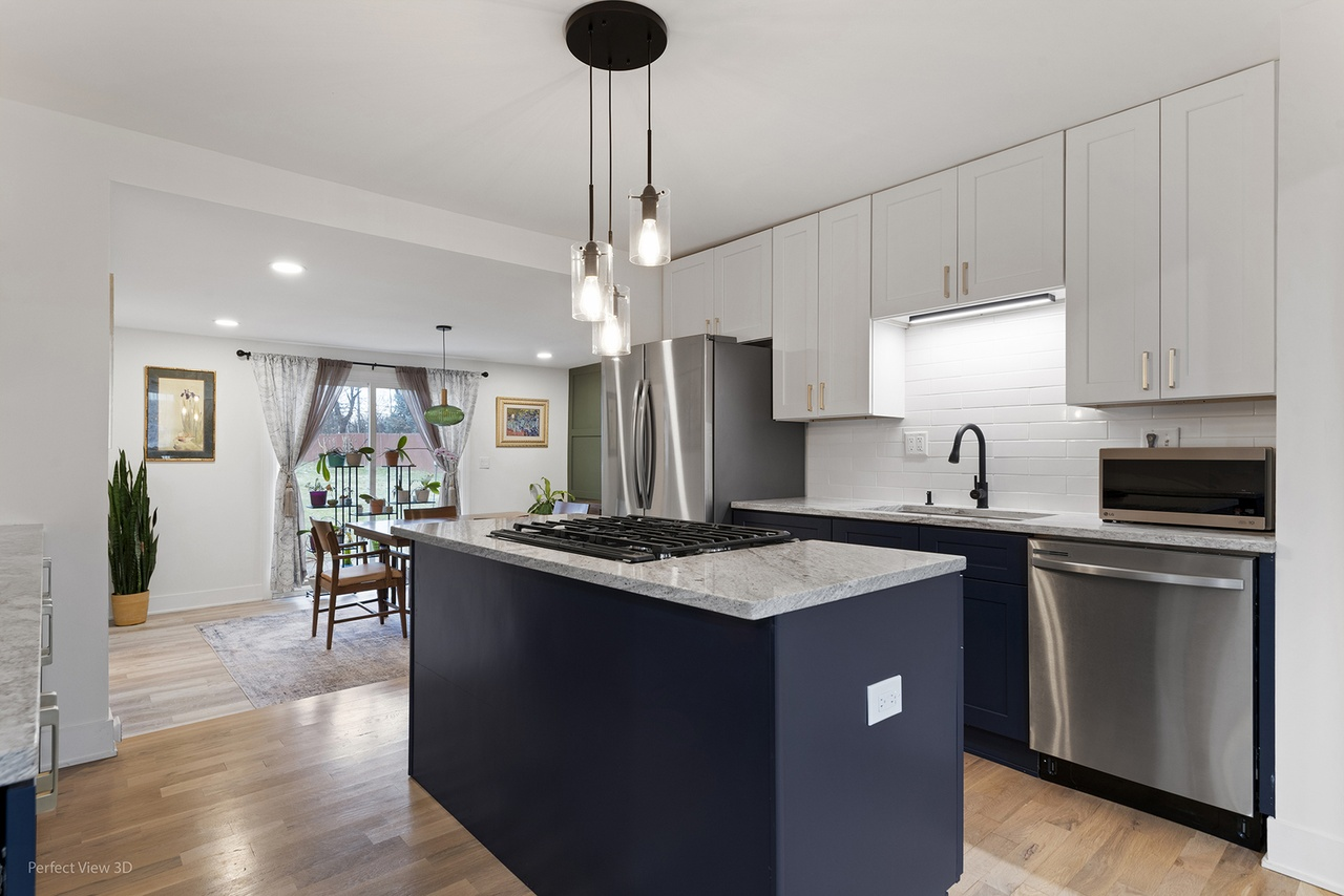 Modern kitchen with white upper cabinets, navy lower cabinets, stainless steel appliances including a refrigerator and dishwasher, a black sink, marble countertops, and pendant lighting. A view of a dining area with a table, chairs, and a window with