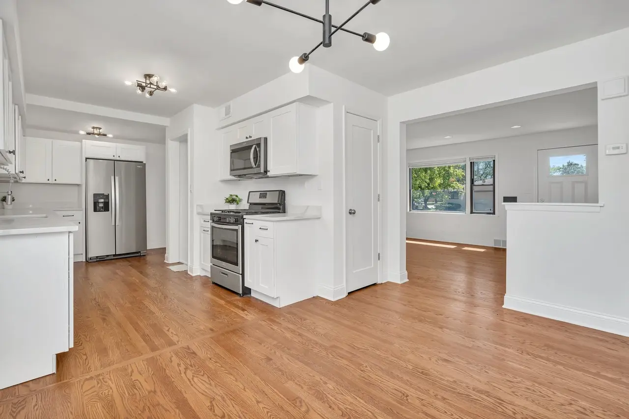 Clean modern kitchen with white cabinets, stainless steel appliances, hardwood floors, and an adjacent living area with large windows letting in natural light.