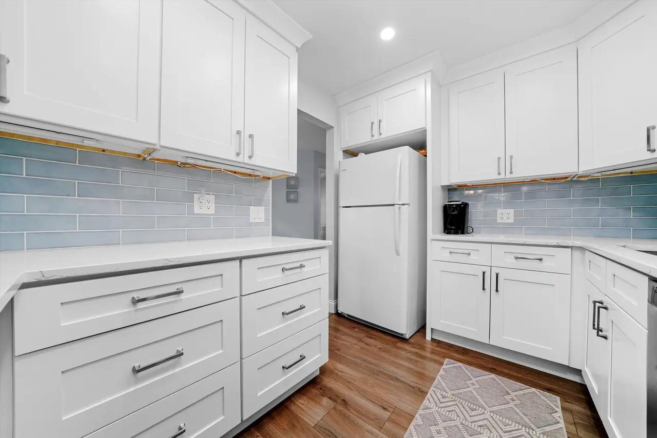White kitchen with white cabinets, light blue tile backsplash, white refrigerator, black coffee maker, wooden floor, small beige rug