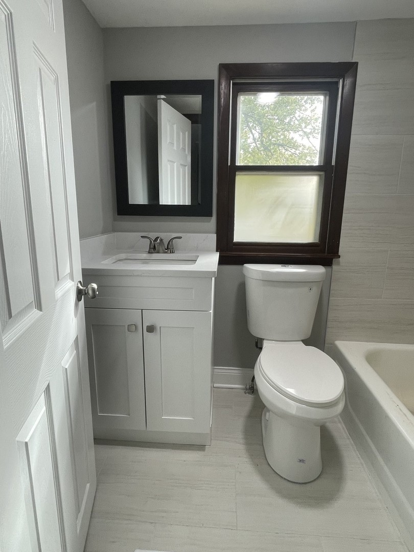 Small bathroom with a white sink vanity, a mirror, a toilet, a window with dark brown trim, and a bathtub, all with light-colored walls and flooring.