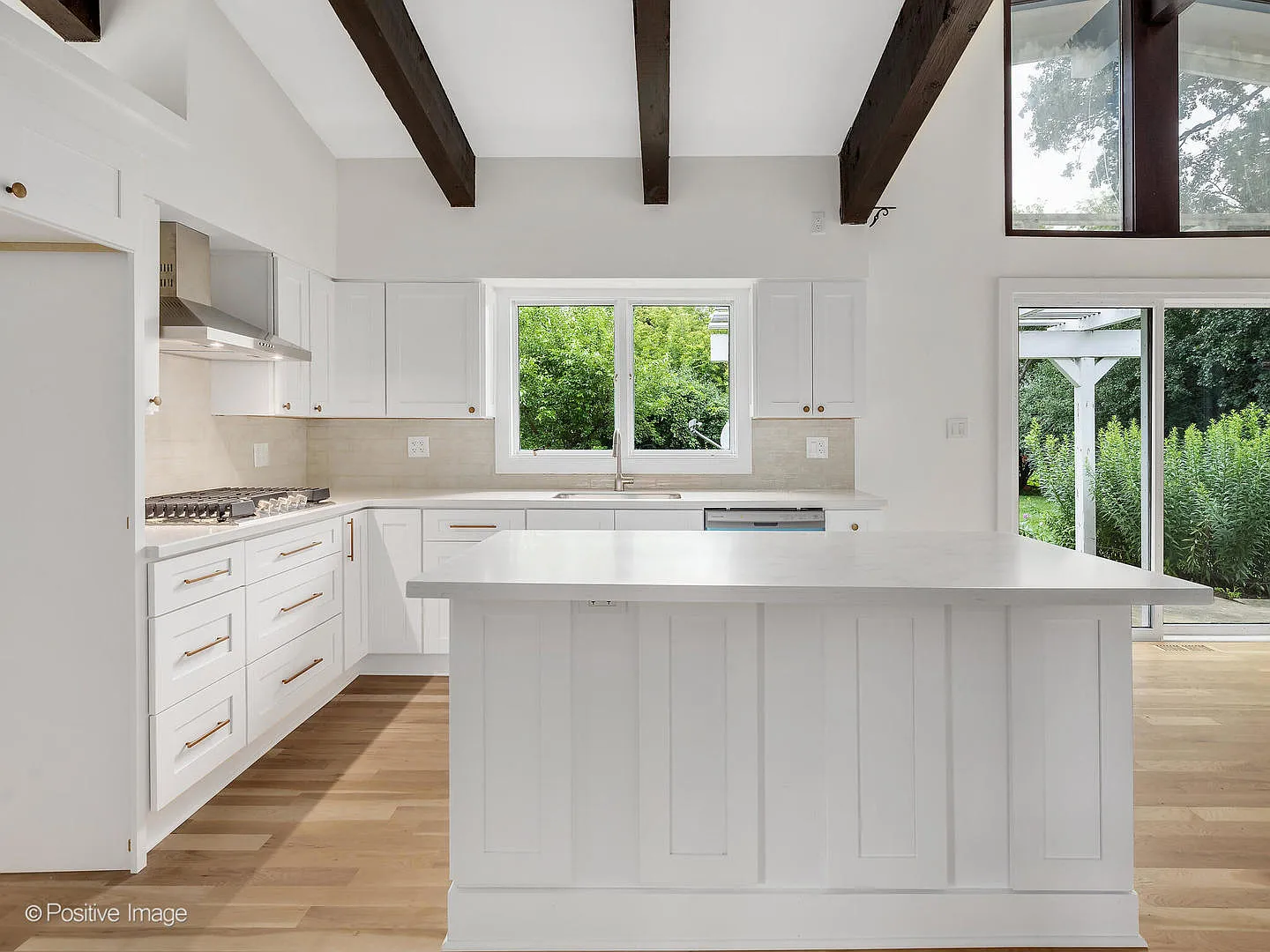 Bright kitchen with white cabinets, wood countertops, a central island, a window over the sink showing greenery outside, and a sliding glass door leading outside. Exposed dark wooden ceiling beams and hardwood flooring.