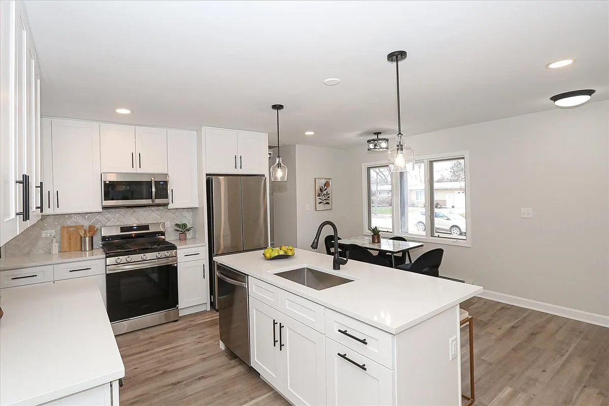Modern open-concept kitchen with white cabinets, stainless steel appliances, including stove, microwave, refrigerator, and dishwasher. Features a white island with a black faucet and sink, pendant lights hanging above, and a dining area with a table 