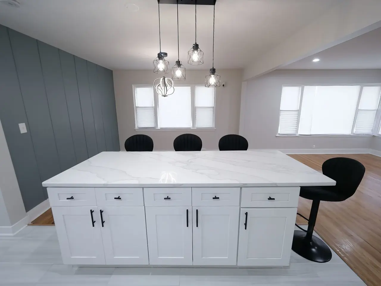 Modern kitchen with a white marble island, black bar stools, pendant lighting, a gray accent wall, and large windows with blinds.