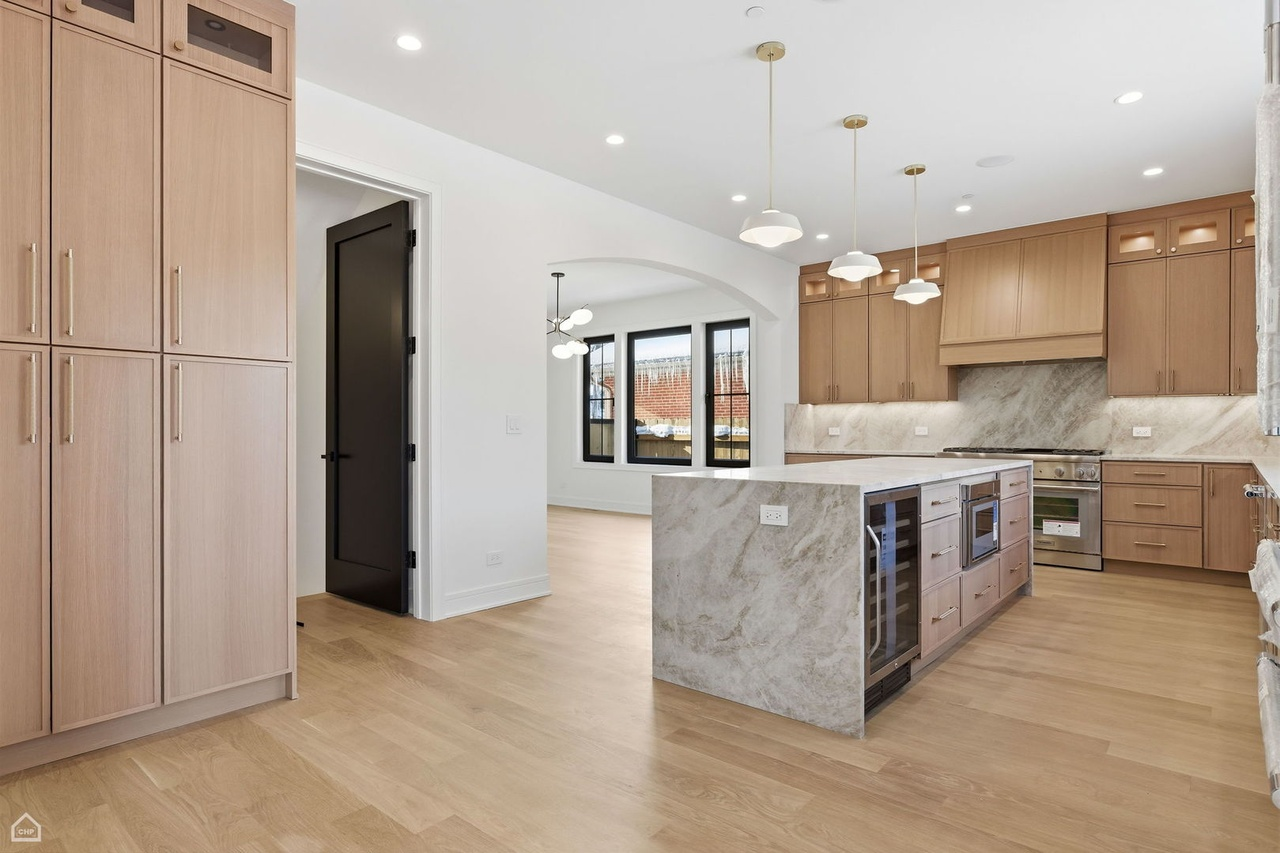 Modern kitchen with wooden cabinets, marble countertops, and a kitchen island. There are multiple pendant lights hanging above the island. The space is open with a small dining area visible through a doorway leading to a room with large windows.
