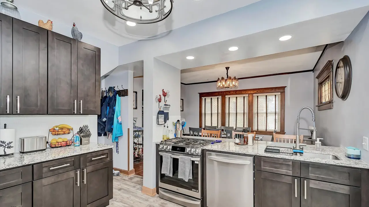Kitchen with dark gray cabinets, granite countertops, and stainless steel appliances. There is a dining area with a chandelier and windows with wooden trim in the background.