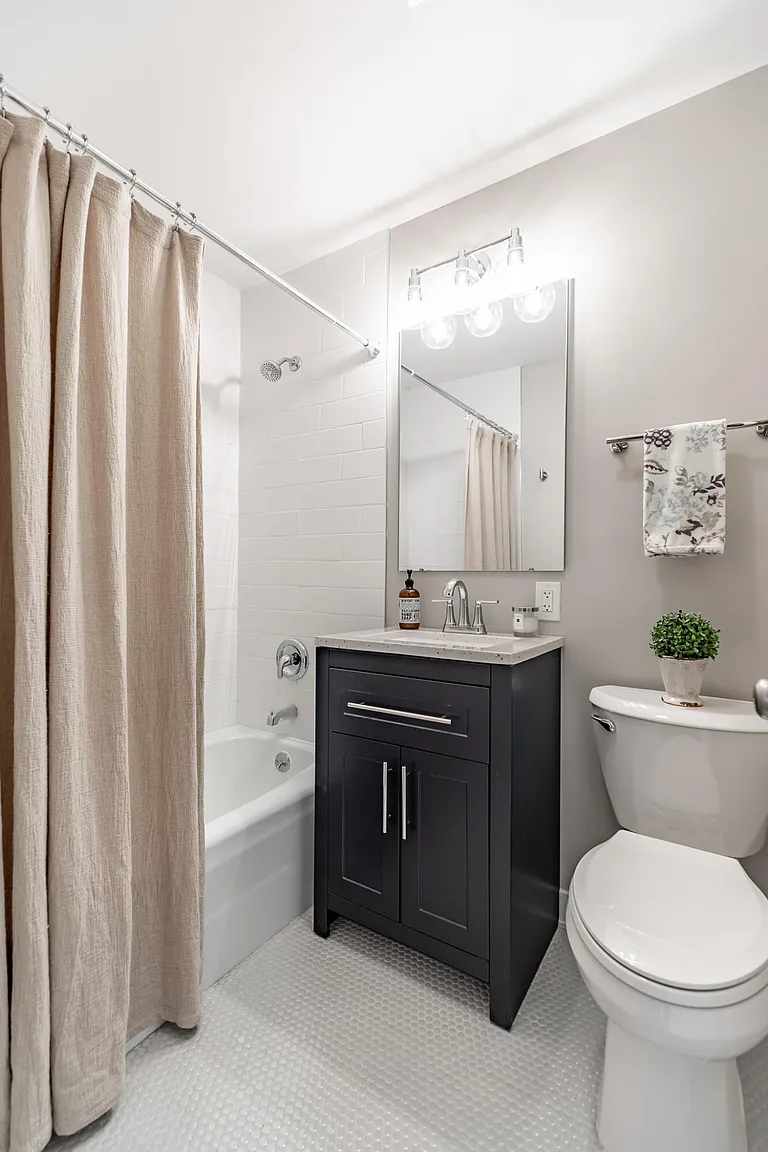 A small bathroom with a beige shower curtain, a white bathtub, a dark vanity with a light countertop, a modern mirror, a bottle of soap, a small potted plant on the toilet, and a decorative towel on a towel rack.