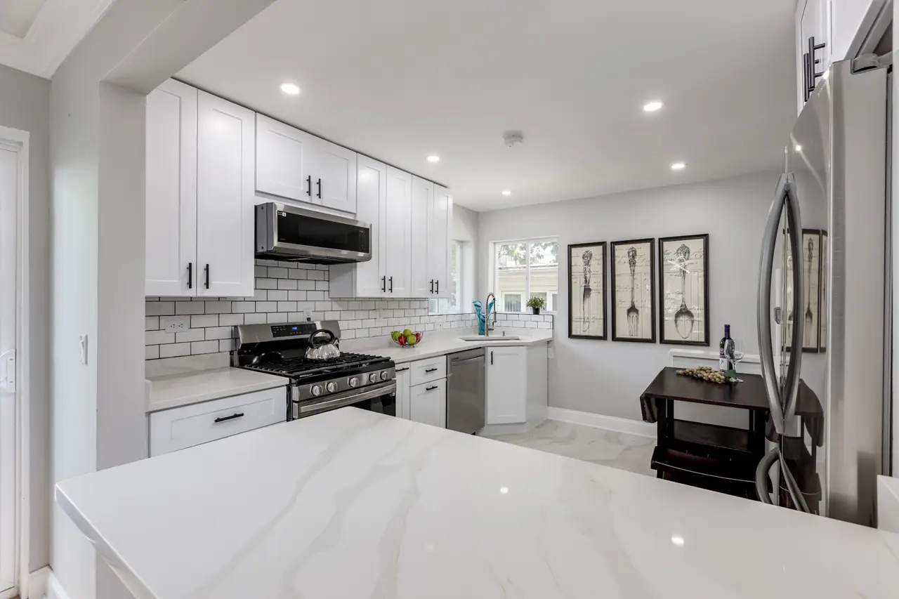 Modern kitchen with white cabinets, stainless steel stove and microwave, white countertop, and a small dining table against the wall featuring three framed skeleton art prints.