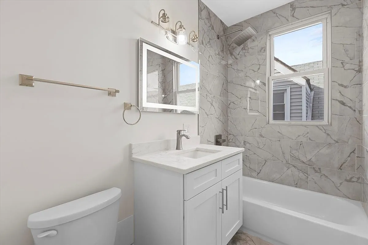 A modern bathroom with a bathtub, a vanity with a sink and mirror, a window, and marble tile walls.