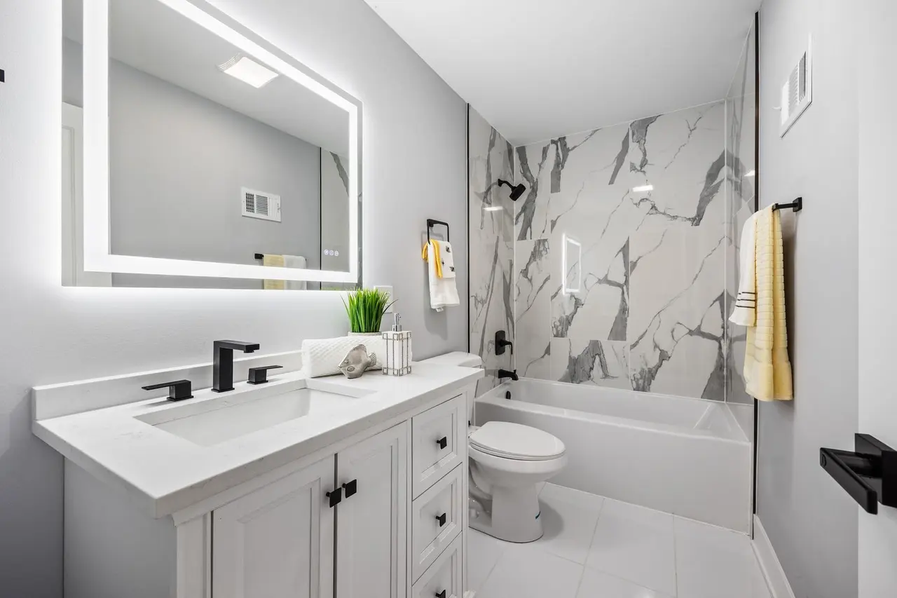 Modern bathroom with white vanity, black fixtures, marble accent wall in the shower, a large backlit mirror, white tiles, and yellow towels.