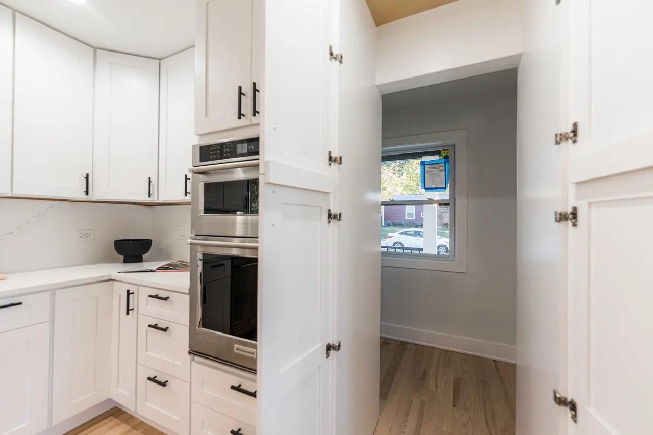 Empty kitchen with white cabinets, black handles, and stainless steel double oven, with a doorway leading to a small room with a window showing a residential neighborhood.