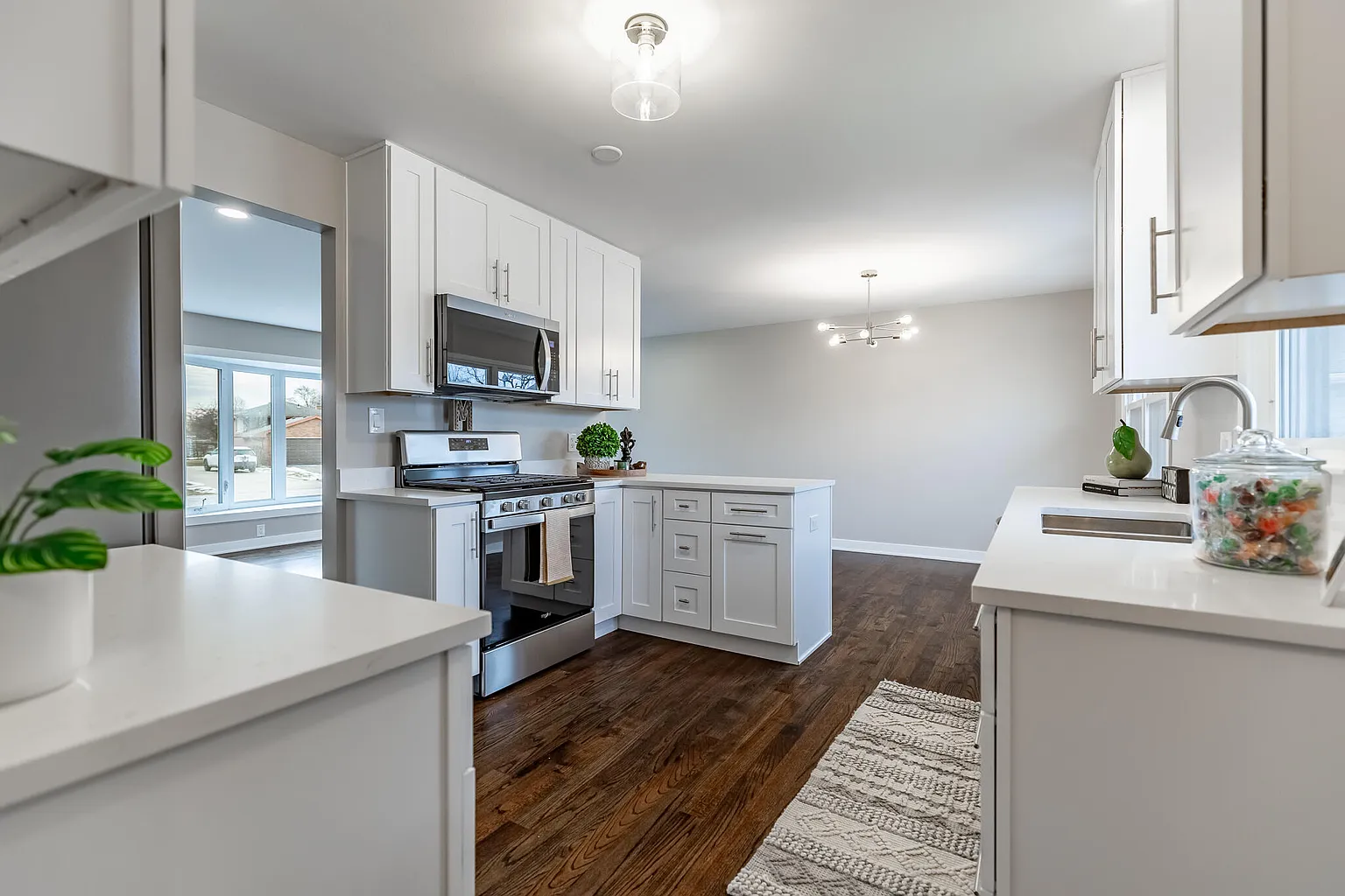 Modern white kitchen with dark wooden floors, stainless steel stove and microwave, white cabinets, and a window with natural light.