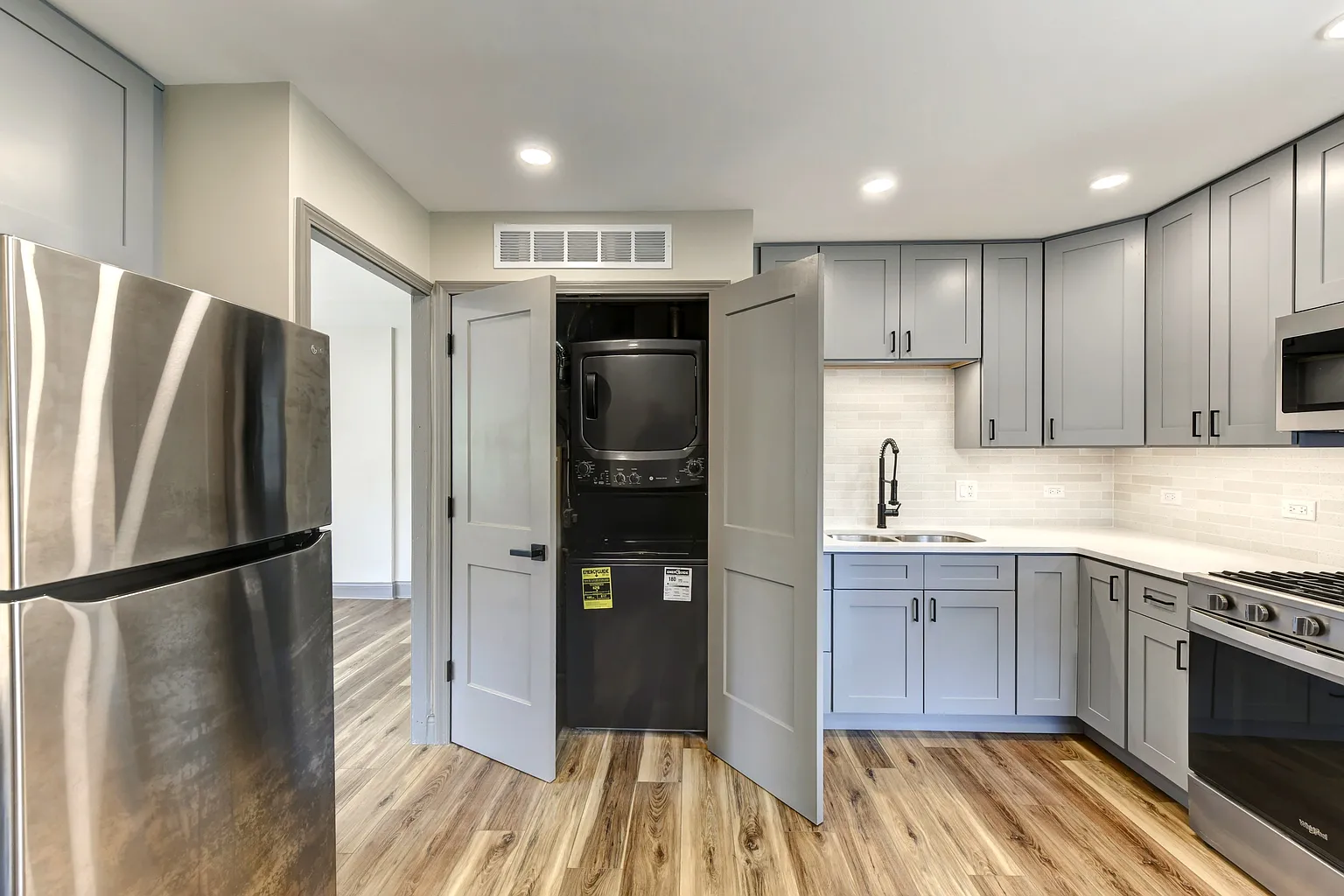 Kitchen with gray cabinets, stainless steel refrigerator, black stove, white subway tile backsplash, and wood-like flooring. A stacked washer and dryer are housed in a closet with open doors.