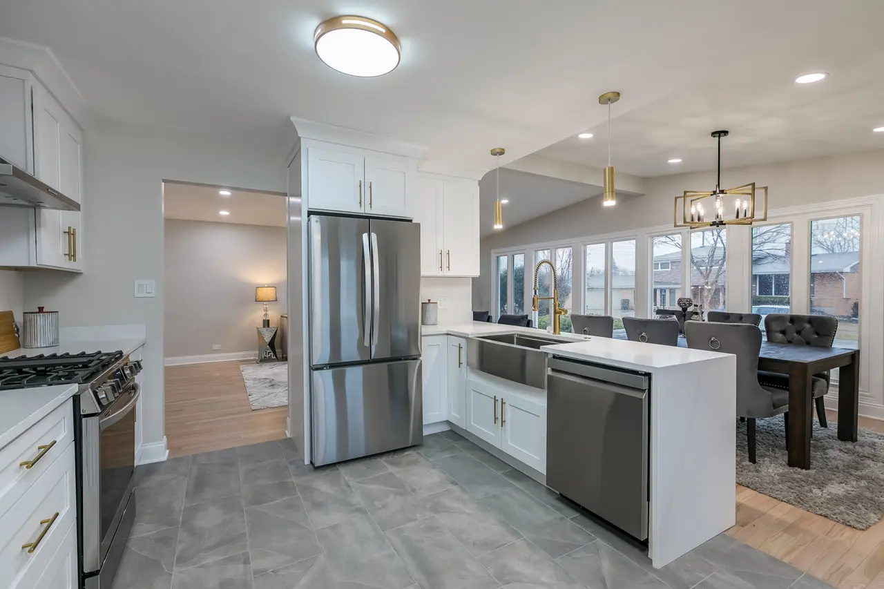 Modern kitchen with white cabinets, stainless steel appliances, gray tiled flooring, and a view into a dining area with large windows and a view outside.