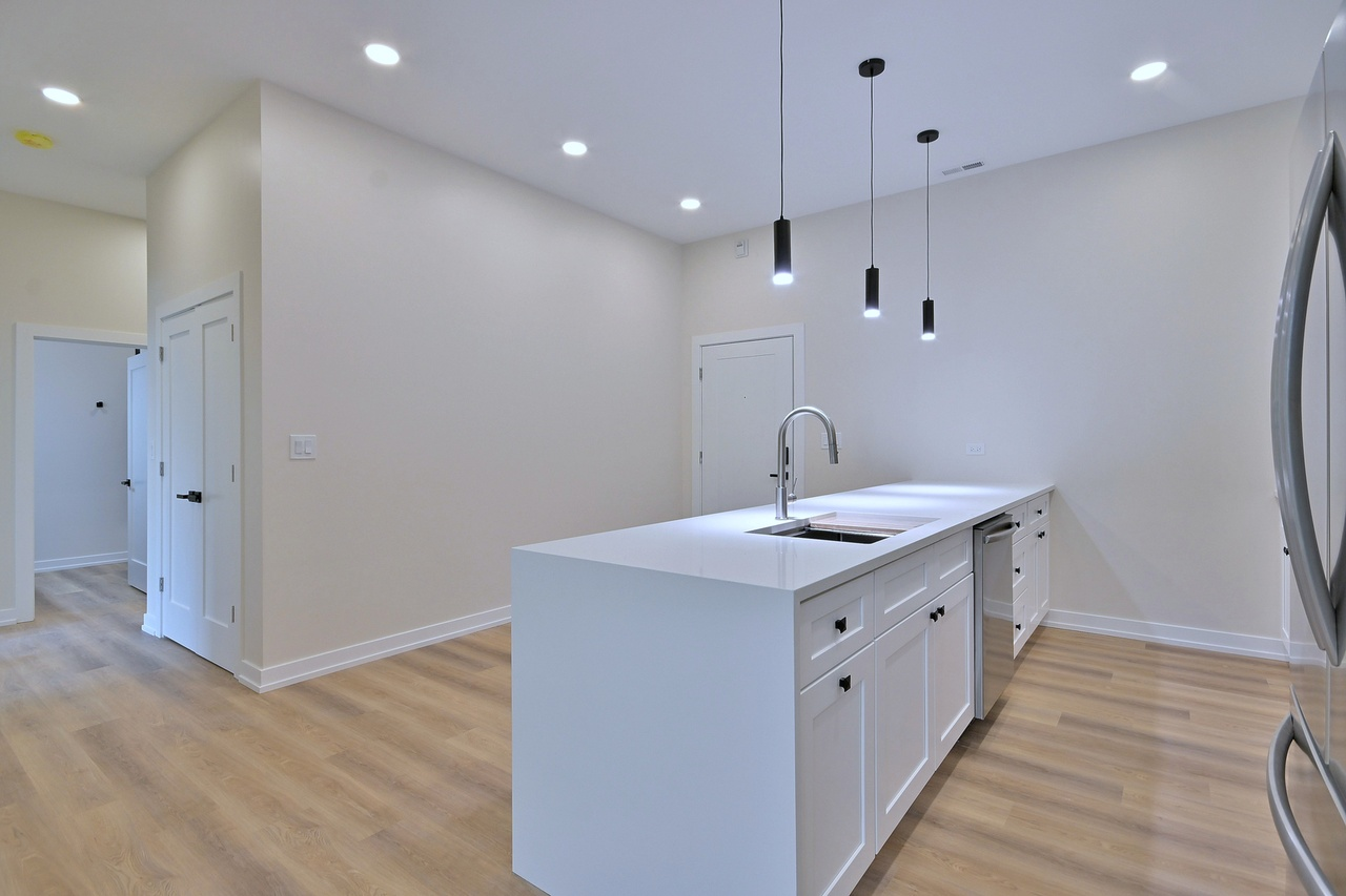 Modern kitchen with white cabinets, a white island with a sink, black pendant lights, and wood flooring.