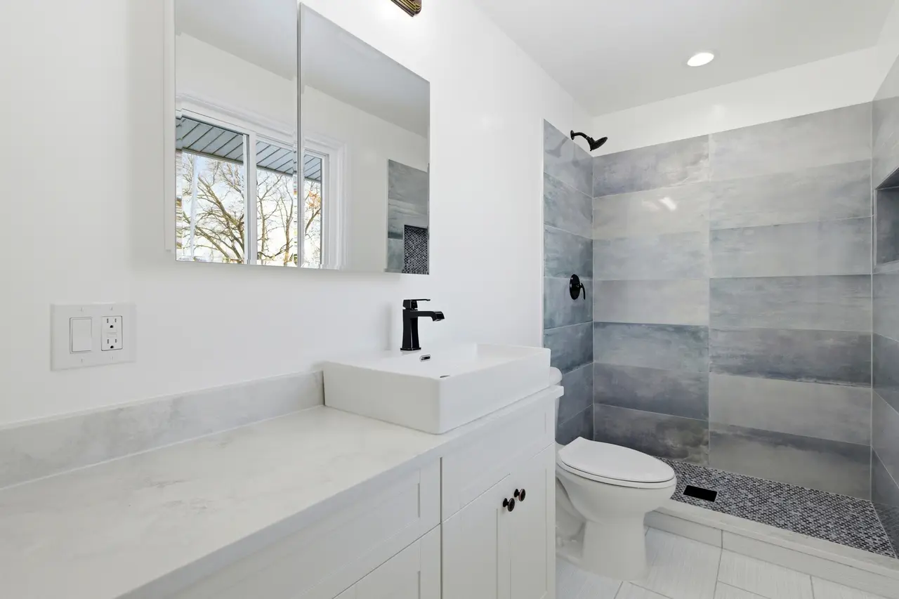 Modern bathroom with white vanity, black faucet, mirror, toilet, and walk-in shower with gray tiles and pebble floor.