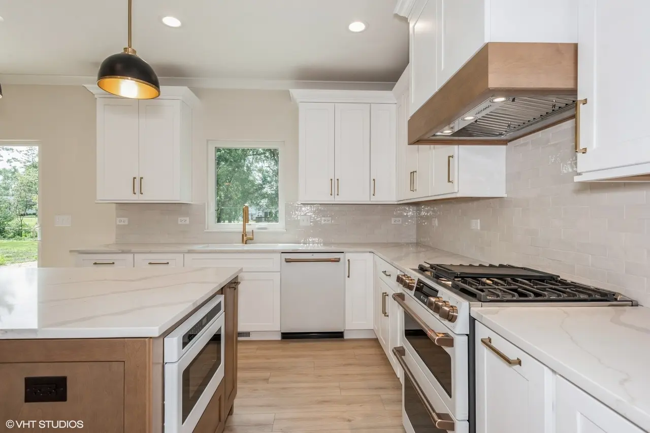 Modern kitchen with white cabinets, marble countertops, a window over the sink, and stainless steel appliances including a stove, oven, and dishwasher. There is a black and gold hanging light fixture and wooden flooring.