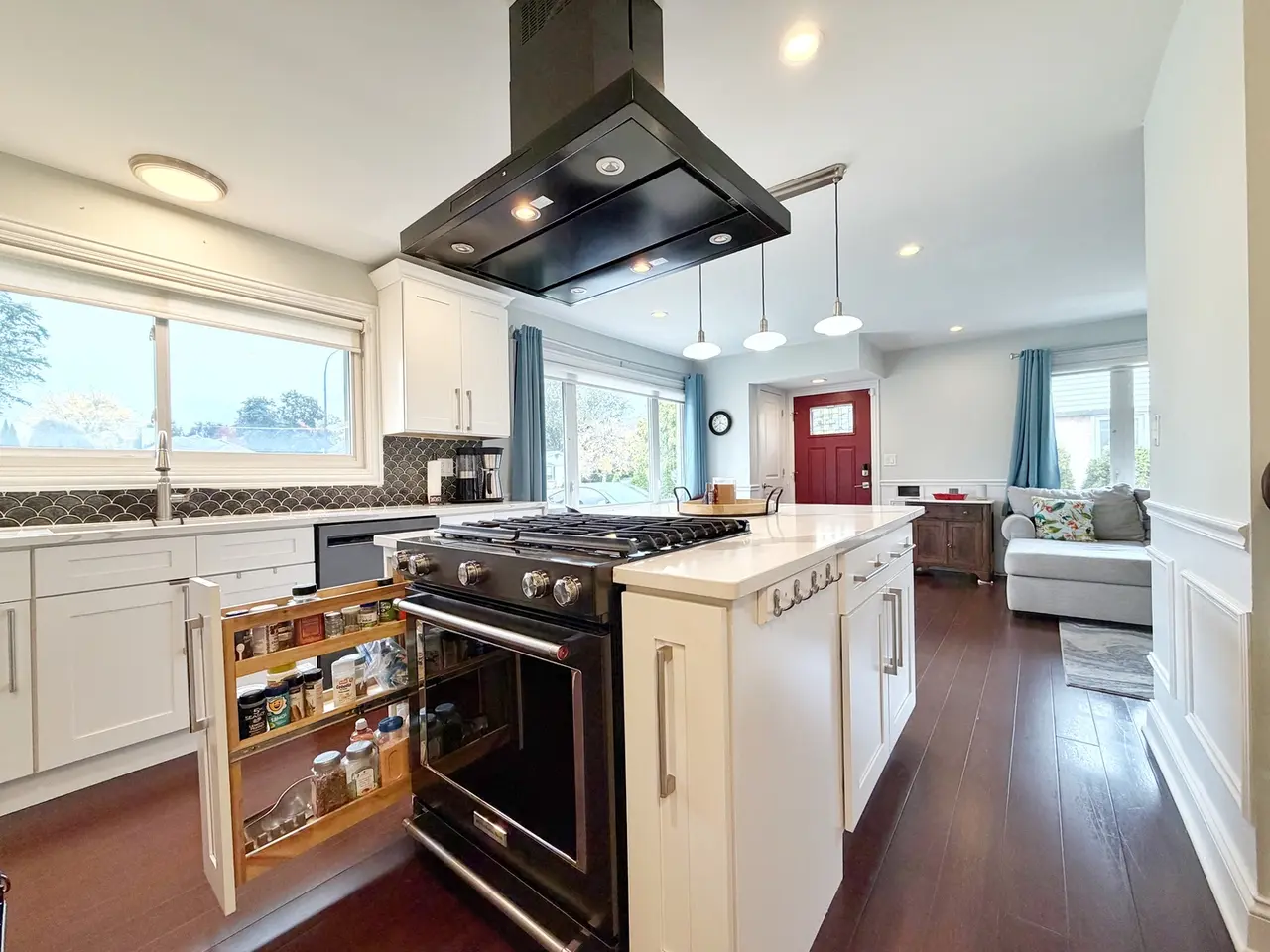 Modern kitchen with white cabinets, black stove, open pantry with spices, large window, and view of living area with red front door and gray sofa.