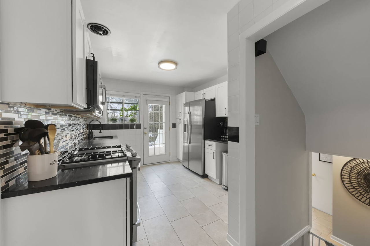 Modern kitchen with white cabinets, black countertops, stainless steel appliances, mosaic tile backsplash, and a door leading outside.