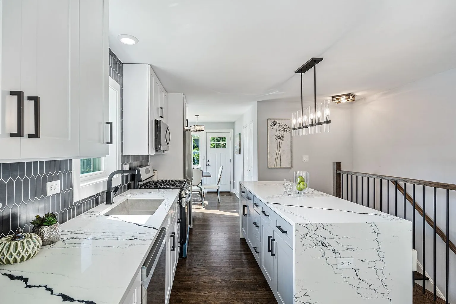 Modern kitchen with white cabinetry and dark hardware, black hexagon tile backsplash, white marble countertops with black veins, and a large kitchen island. There are pendant lights above the island and a window over the sink. A small dining area is 