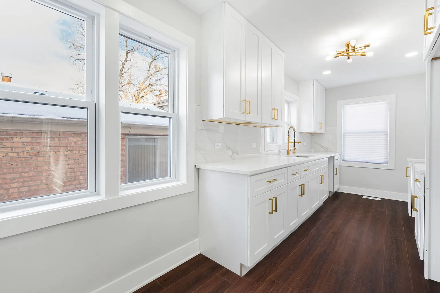 White kitchen with gold accents, dark wood flooring, and large windows, featuring white cabinets, a marble countertop, and a gold faucet.