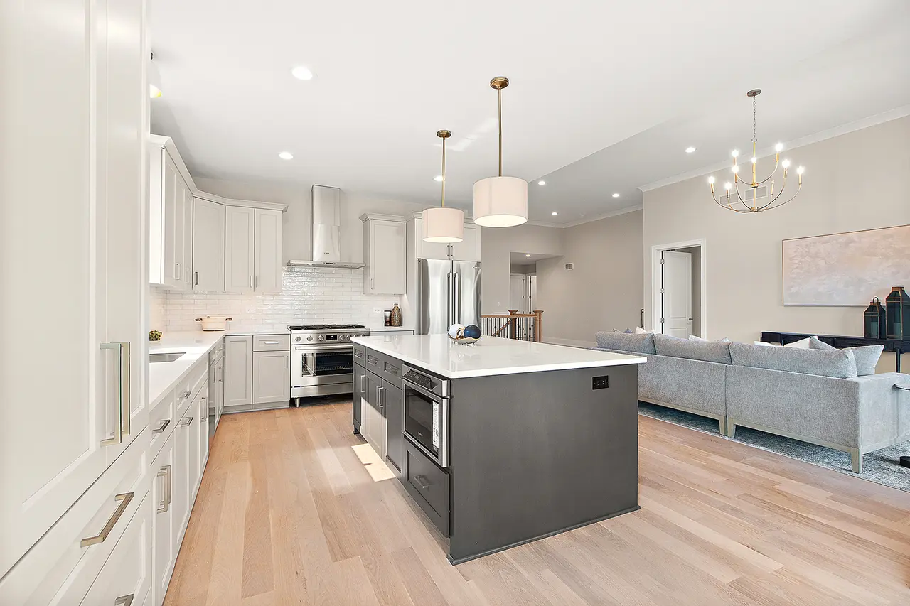 Open-concept modern kitchen and living room with white and gray color scheme, featuring an island with a microwave, kitchen appliances, light fixtures, and a gray sofa.