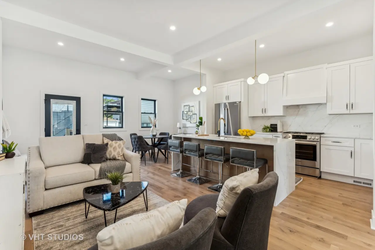 Open-concept kitchen and living room with white cabinets, stainless steel appliances, black barstools at a kitchen island, beige sofa with throw pillows, dark armchair, dining table with chairs, hardwood floors, and modern lighting fixtures.