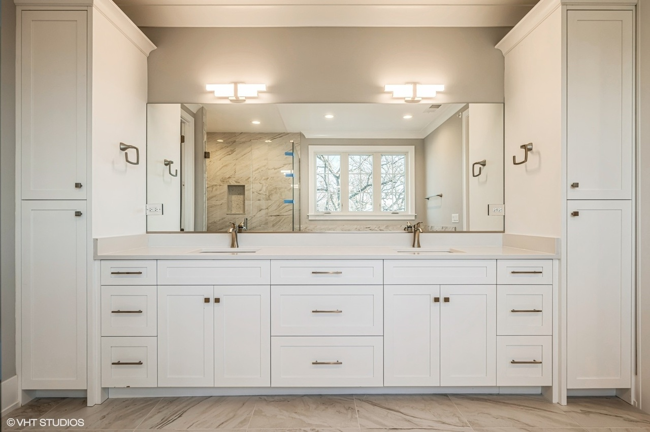 A modern bathroom vanity with white cabinets, a large mirror, and two sinks with gold faucets, illuminated by two lights on the wall above.