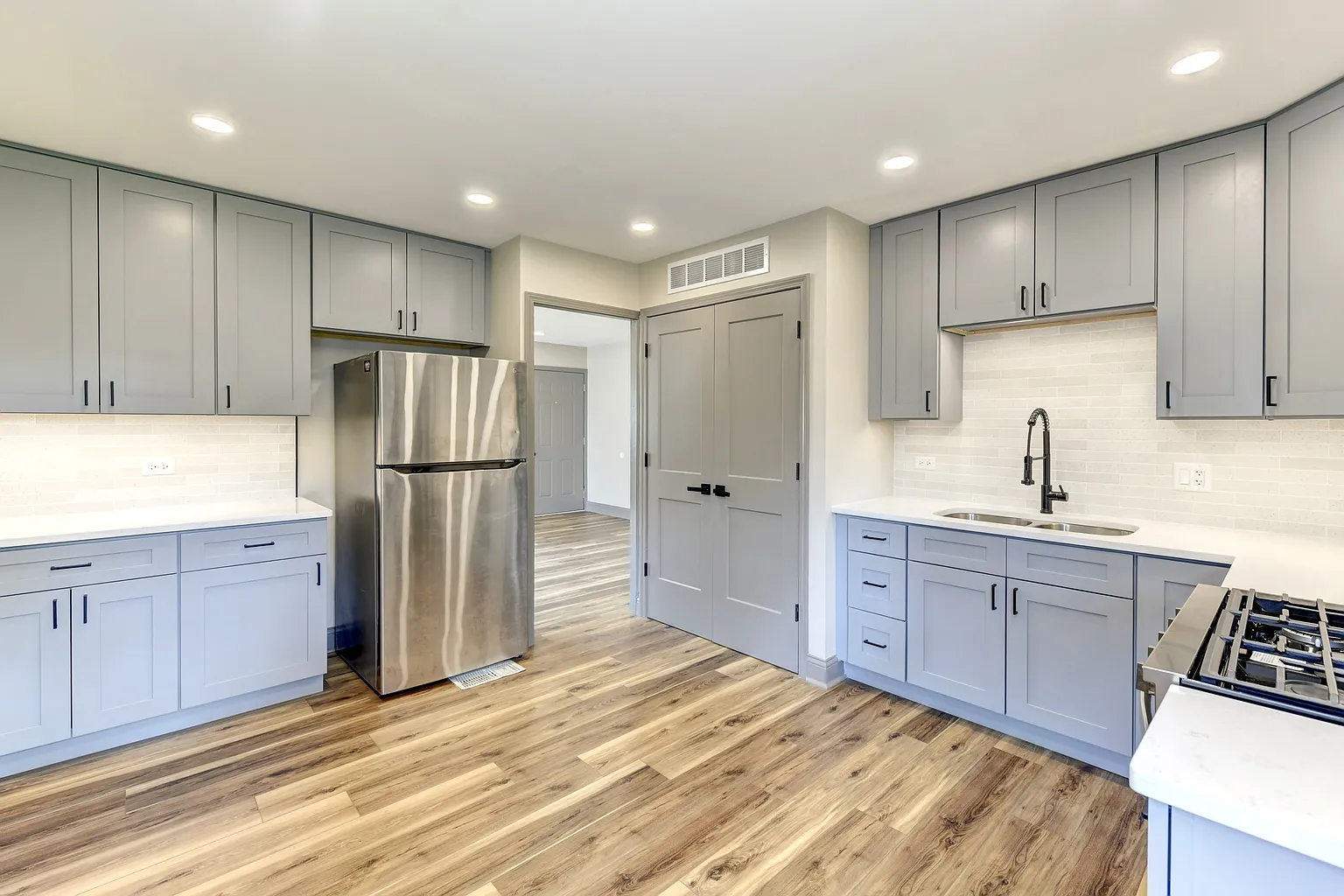 Modern kitchen with light gray cabinets, stainless steel refrigerator, white countertop, white brick backsplash, black faucet, and wood-look flooring.