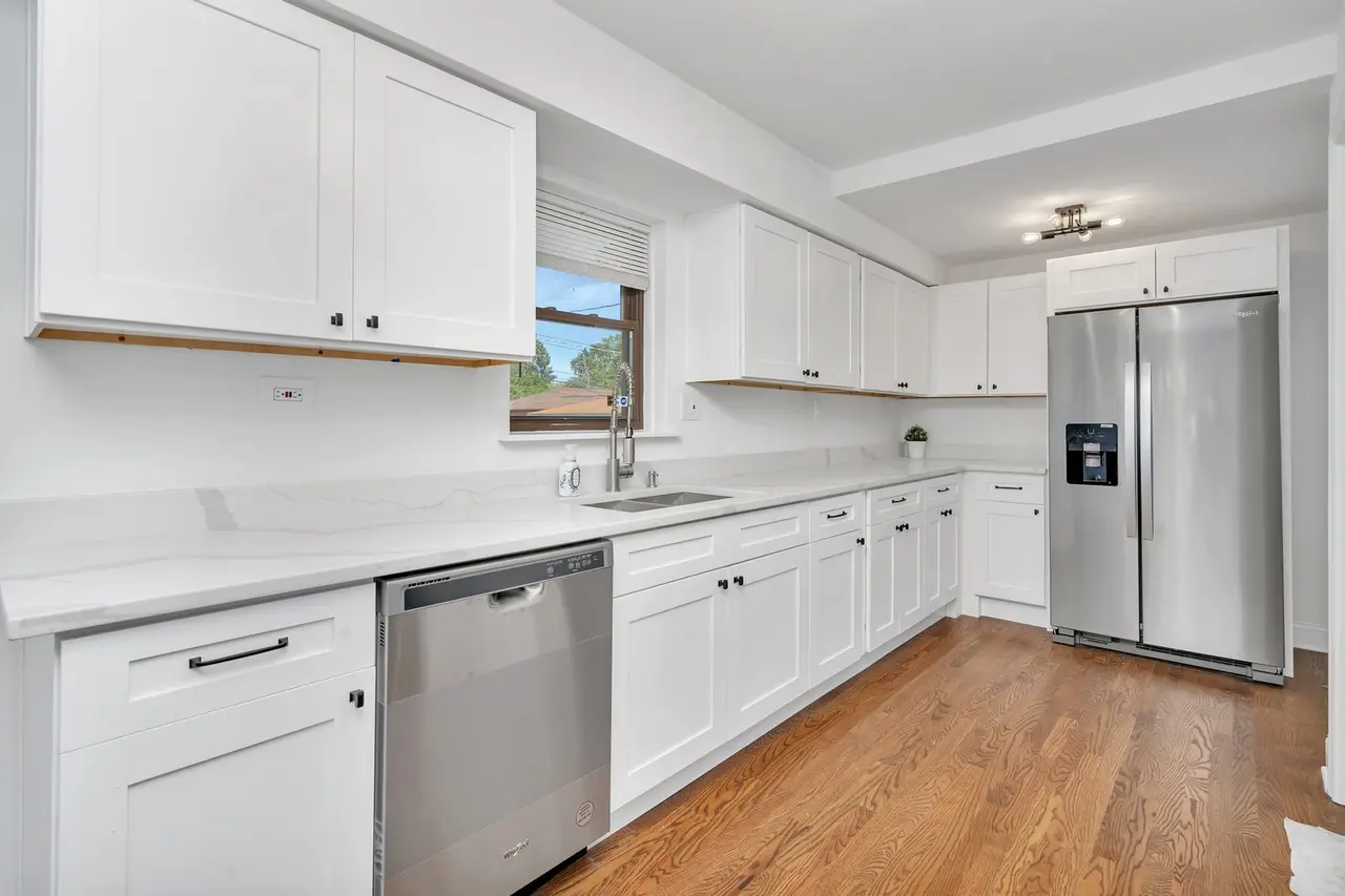 Kitchen with white cabinets, stainless steel refrigerator, dishwasher, small plant, window, and wooden flooring.