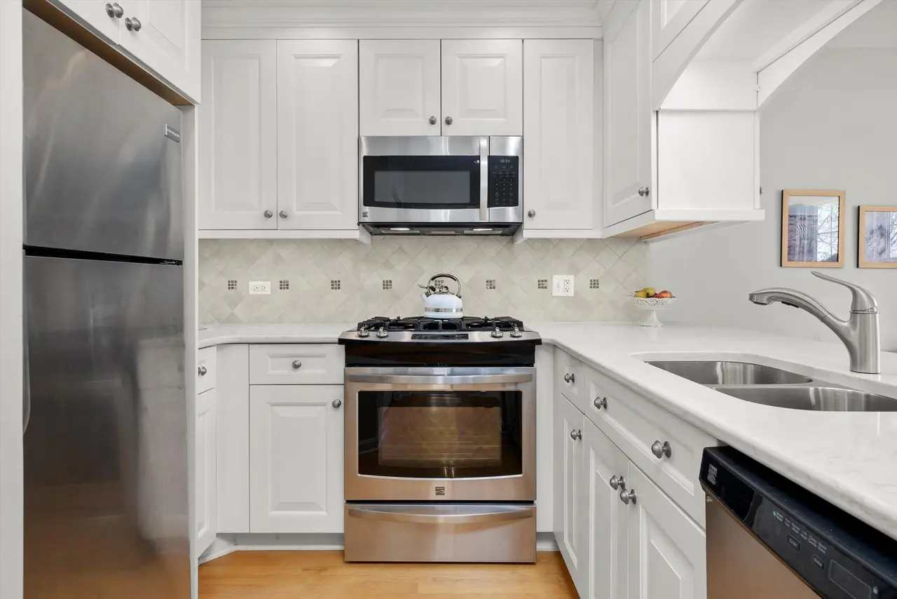 Modern kitchen with stainless steel refrigerator, microwave oven, gas stove, and double sink. White cabinetry and beige tile backsplash.