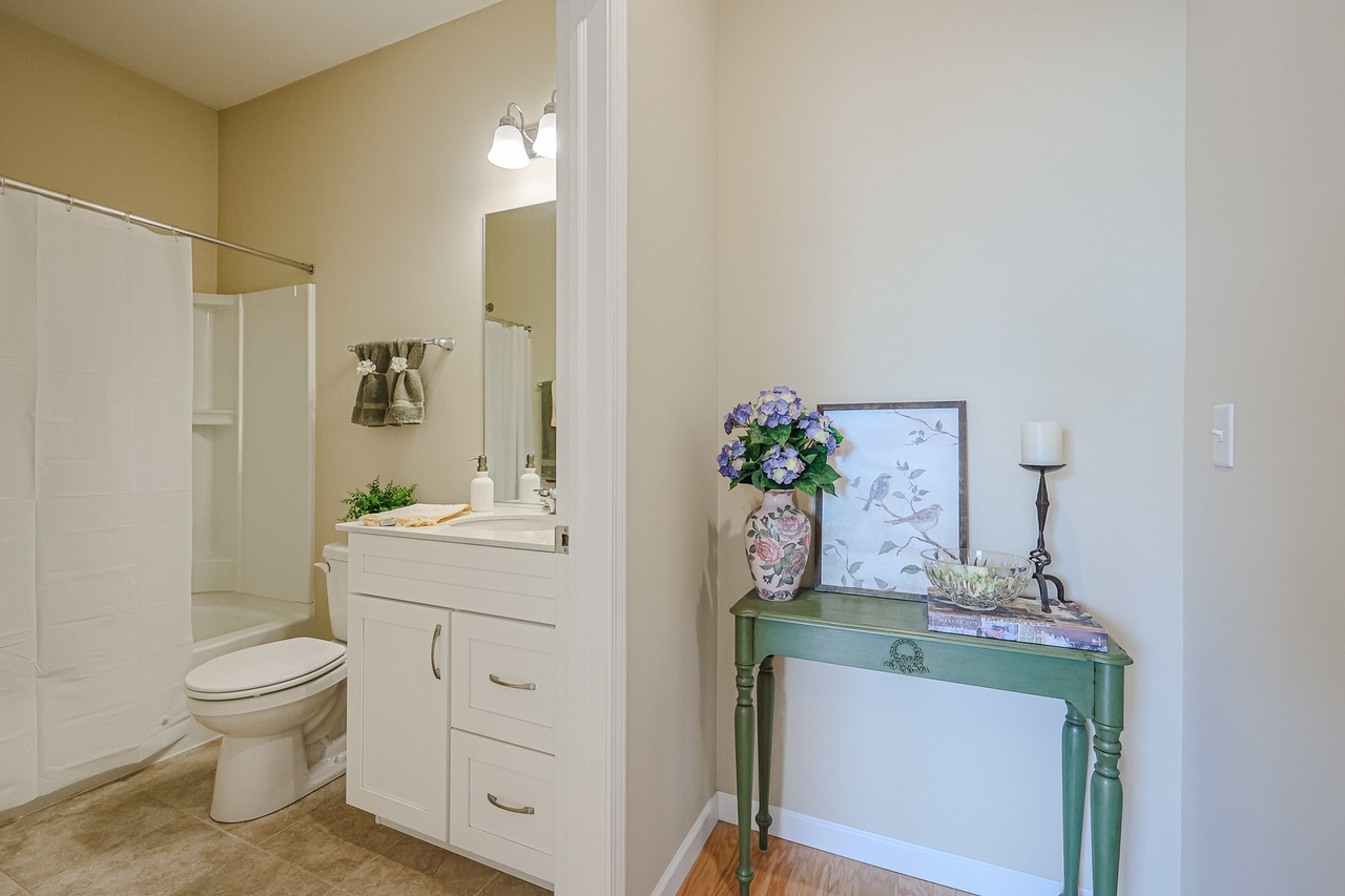 Bathroom with shower, toilet, mirror, sink, towel, and decorative table with vase, framed picture, candle, and bowl.