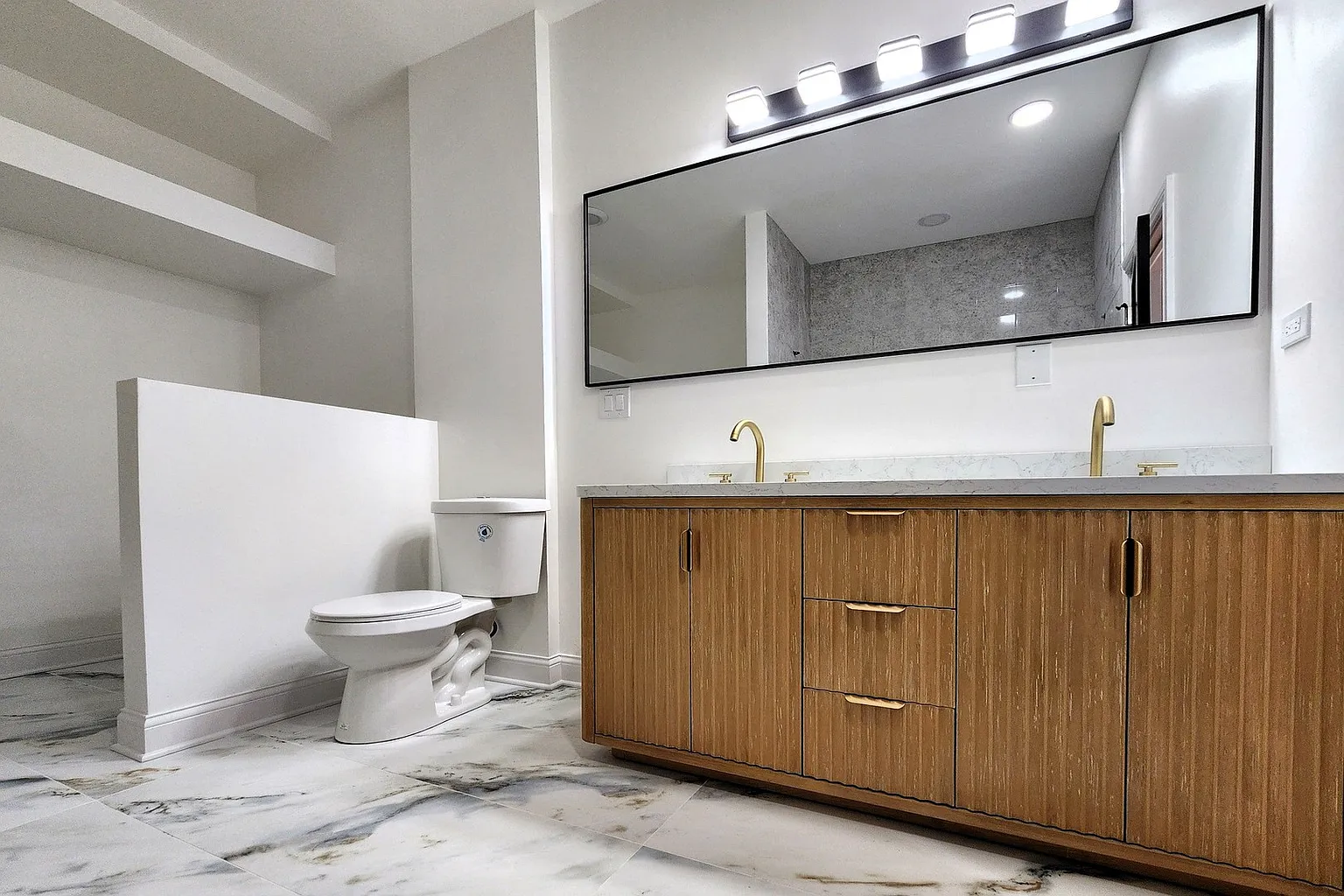 Modern bathroom with marble flooring, white walls, a wooden vanity with two gold faucets, a large mirror, and a toilet.
