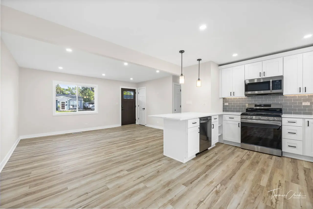 Modern kitchen with white cabinets, stainless steel appliances, gray subway tile backsplash, pendant lighting, hardwood flooring, and an open living space with a window showing a view of a sunny neighborhood.