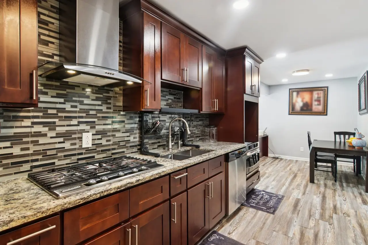 Kitchen with dark wood cabinets, granite countertops, a stainless steel range hood, and a mosaic tile backsplash, leading to a dining area with a table, chairs, and framed artwork on the light-colored wall.