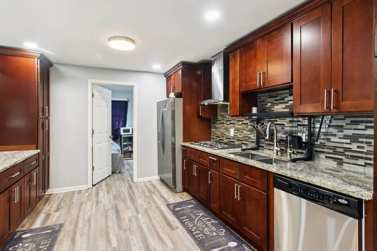 Kitchen with dark wood cabinets, granite countertops, mosaic tile backsplash, stainless steel appliances, and hardwood flooring. Visible doorway leading to another room.