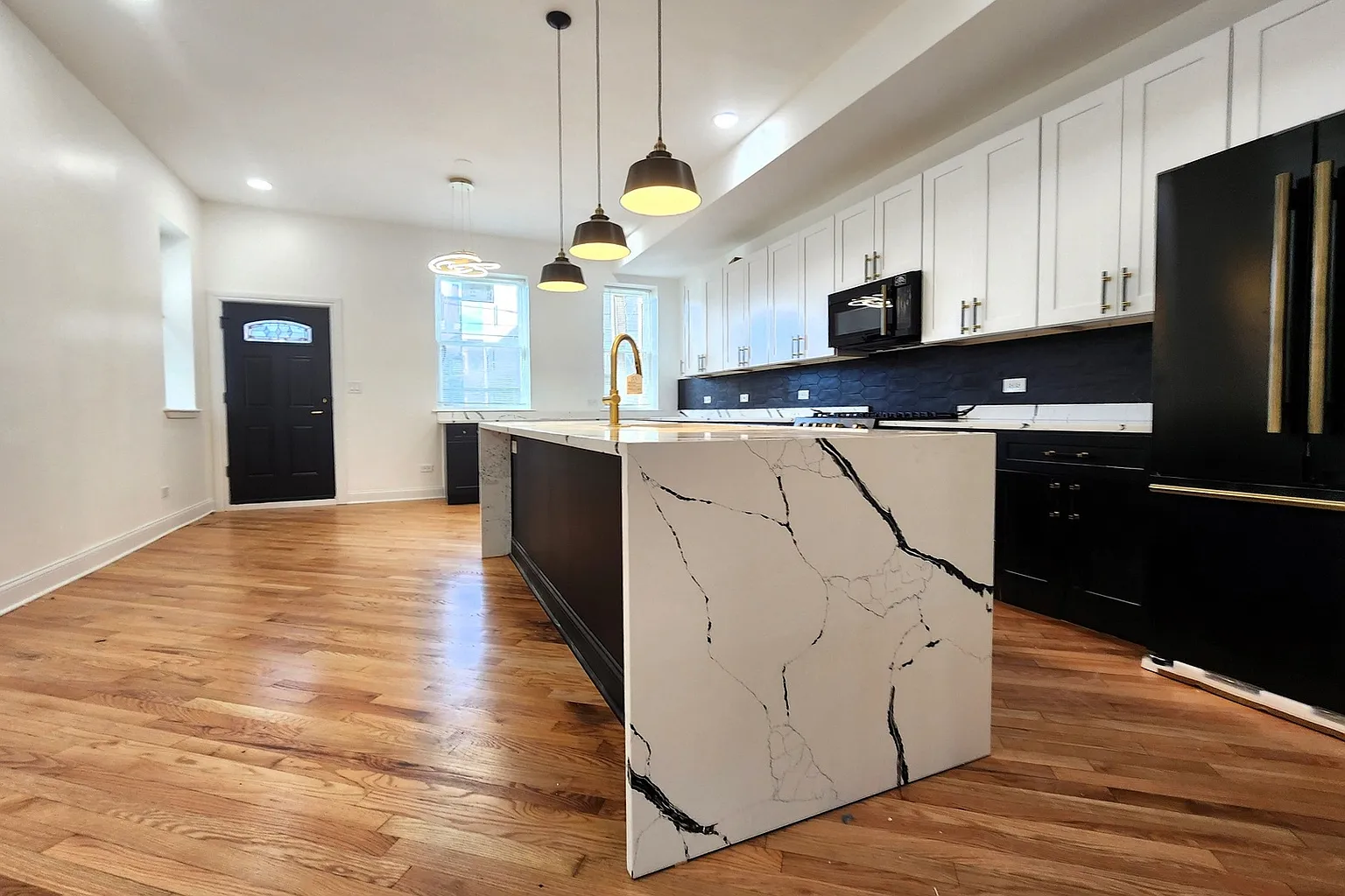 Modern kitchen with white cabinets, black appliances, and wooden flooring. Features an island with a marble-like countertop and gold faucet, pendant lighting, and a black accent wall behind the stove.