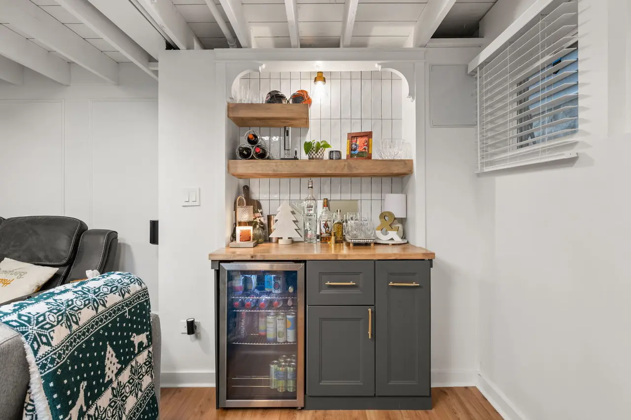 A cozy wet bar area with dark gray cabinets, a wooden countertop, and open shelves decorated for the holidays with a small white Christmas tree, candle, and festive ornaments. There is a mini fridge with drinks, white tiled wall, and a window with wh