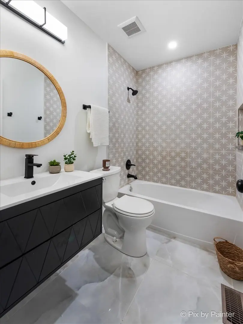 Modern bathroom with a black vanity, white countertop, oval mirror with rattan frame, small potted plants, white toilet, bathtub with star-patterned tile wall, black fixtures, wicker basket, and marble floor.