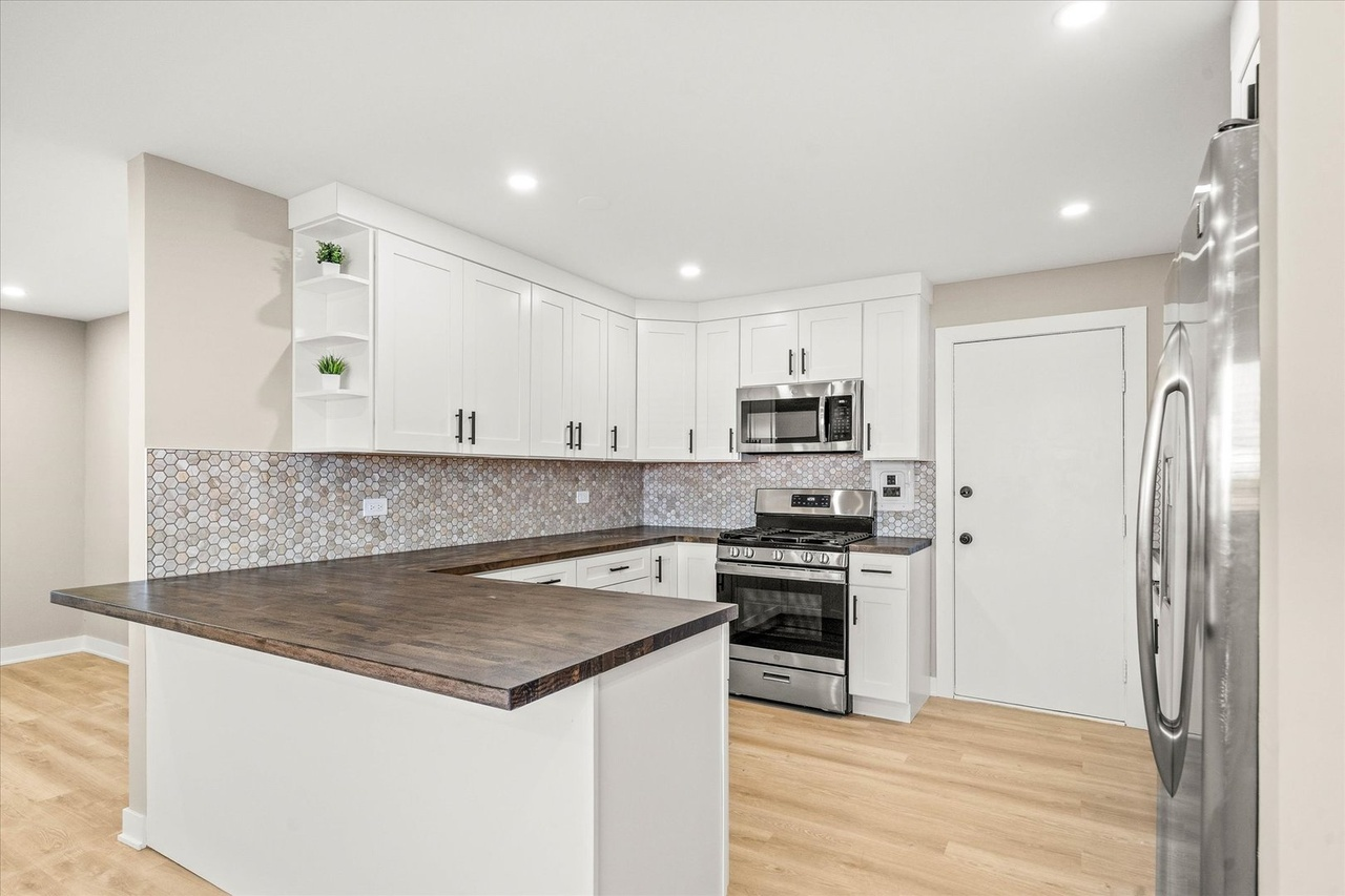 Modern kitchen with white cabinets, stainless steel appliances, wood countertop island, hexagonal tile backsplash, light wood flooring, and recessed ceiling lights.