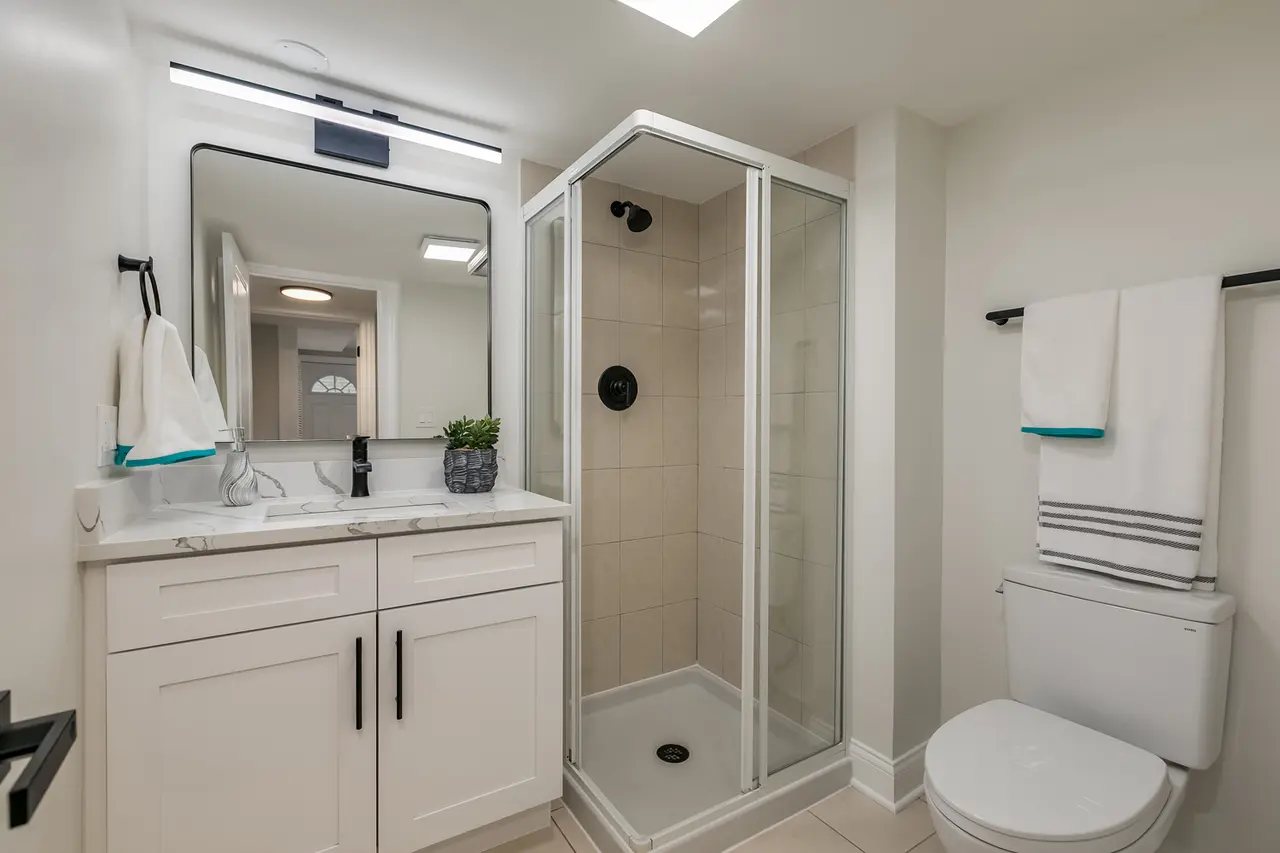Modern bathroom with a white vanity, a large mirror, a small potted plant, a glass soap dispenser, a glass-enclosed shower with beige tiles, a black showerhead, and a white toilet with a towel hanging above it.