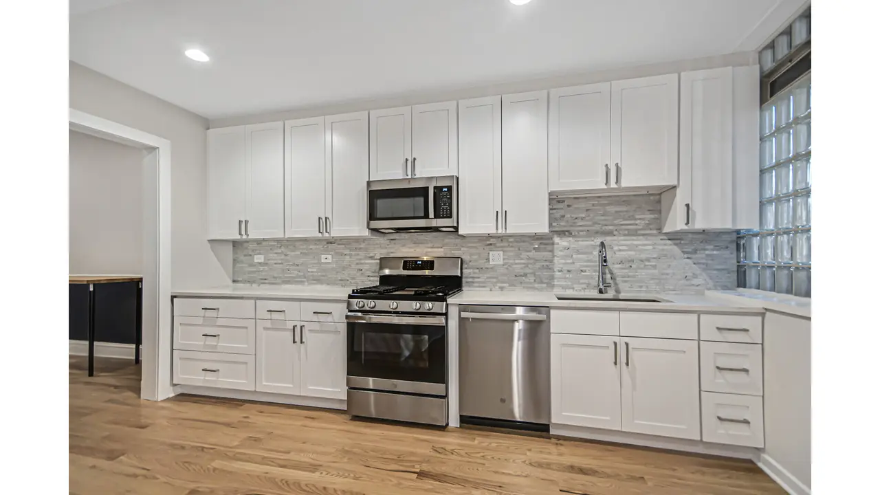 Modern white kitchen with stainless steel appliances, gray backsplash, and hardwood floors.