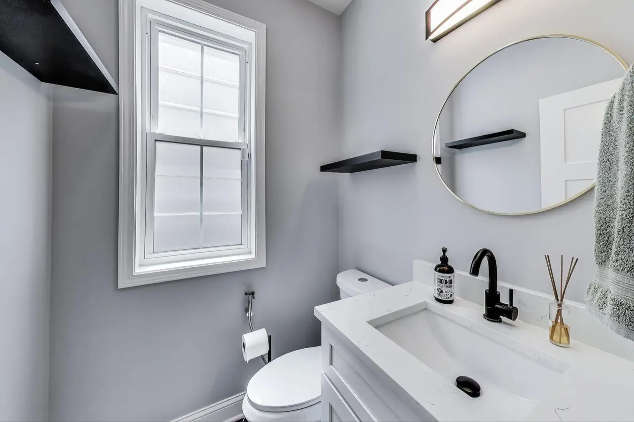 Small modern bathroom with a white sink, black faucet, round mirror, wall-mounted black shelves, gray walls, a window, and a towel hanging on the right side.