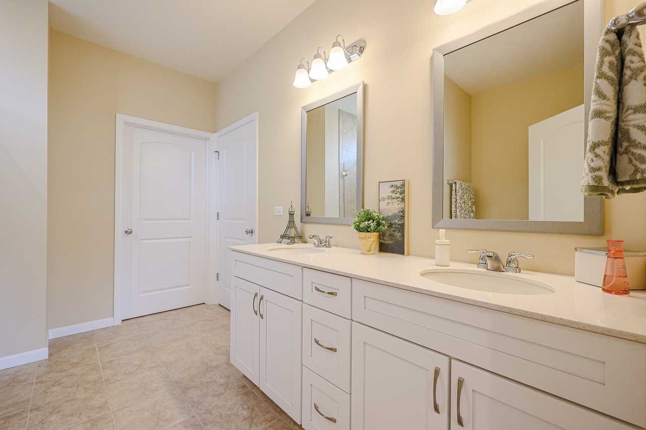 Bathroom with double vanity sinks, large mirrors, beige walls, framed mirrors, and light fixtures, decorated with a small Eiffel Tower figure, potted plant, landscape picture, soap dispensers, and towels.