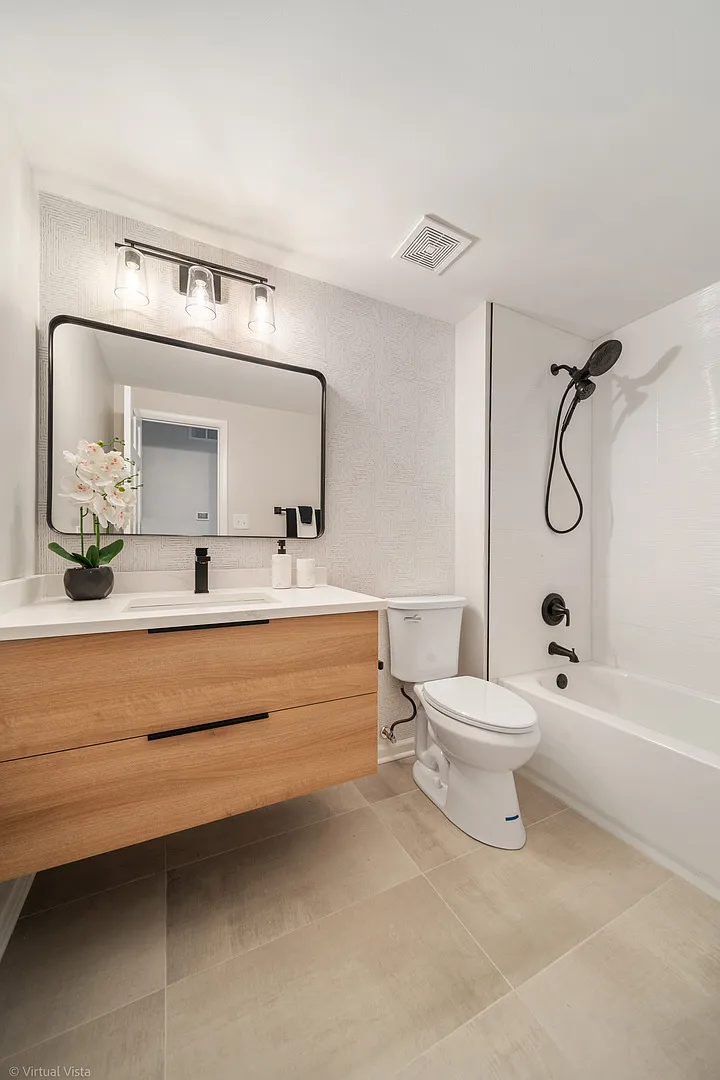 Modern bathroom with wooden vanity, large mirror, potted orchid, toilet, and bathtub with black showerhead and fixtures, beige tile flooring.