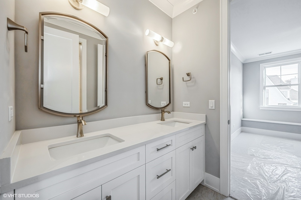 A modern bathroom with double vanity sinks, two oval mirrors, and two wall-mounted light fixtures. The vanity has white cabinetry and brushed nickel handles, with a light gray wall and a doorway leading to an empty room.